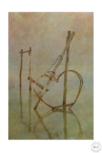 Rusted metal heart structure on wooden posts, partially submerged in calm lagoon water. Fine art photography by RLF Photography