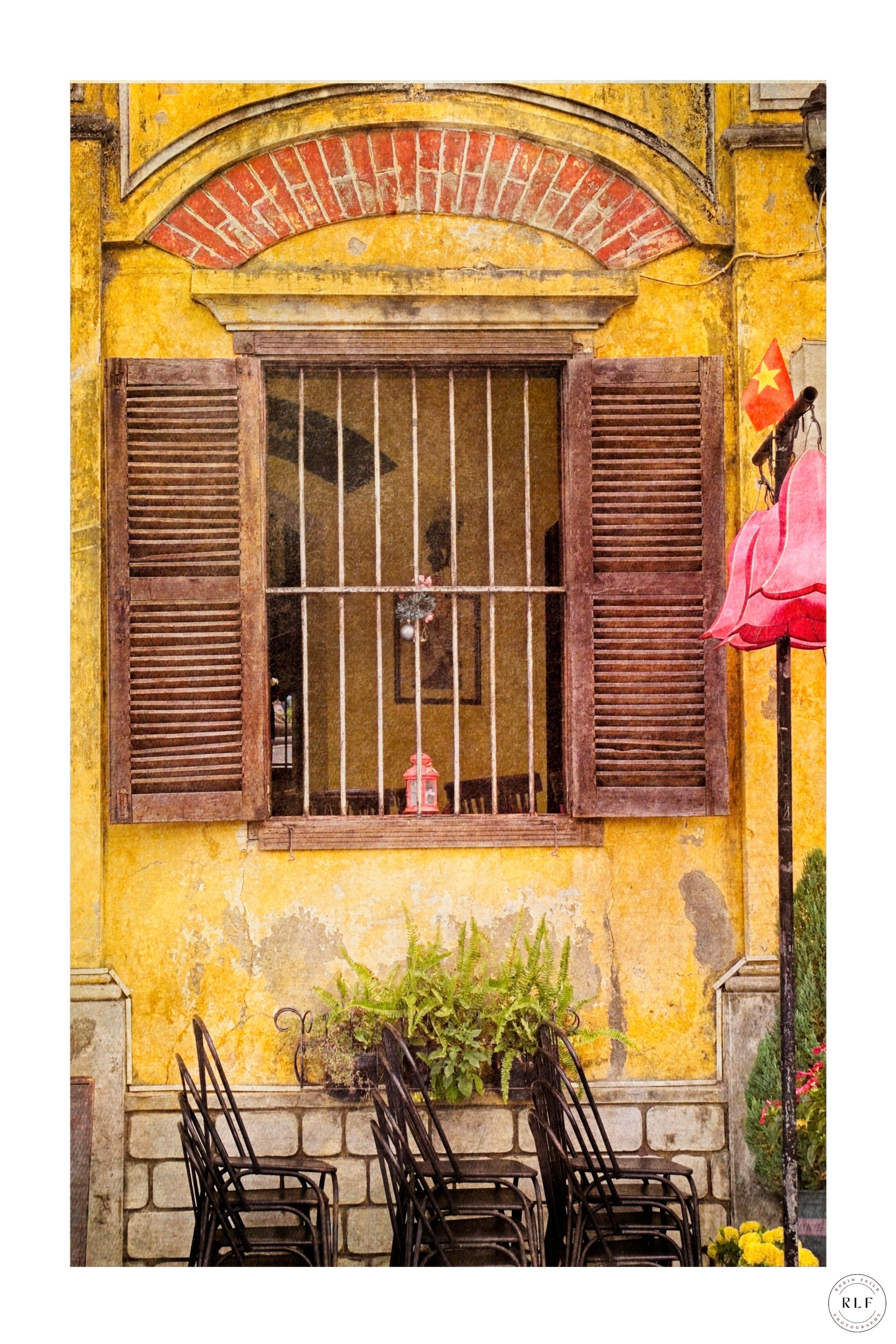 Weathered yellow cafe wall with wooden shutters, metal grille, stacked chairs, and green ferns.