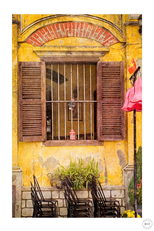 Weathered yellow cafe wall with wooden shutters, metal grille, stacked chairs, and green ferns.